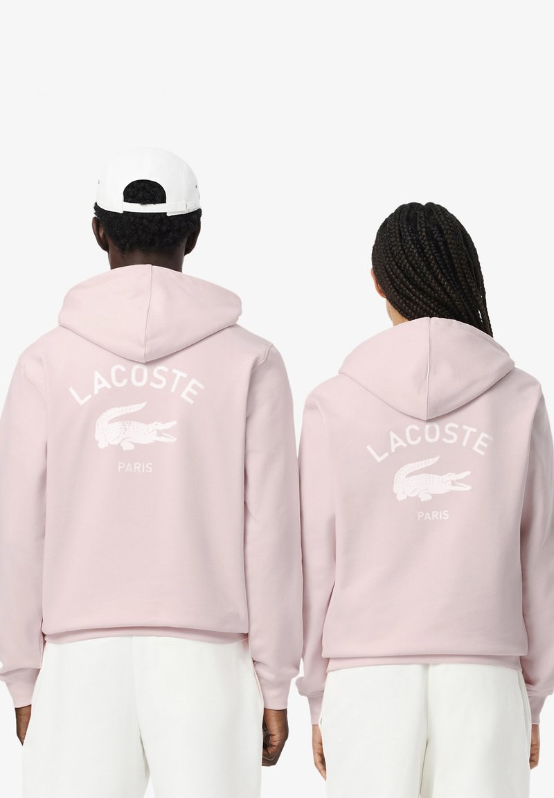 Sudaderas con capucha rosas que presentan el logo en blanco de "LACOSTE" y el cocodrilo en la parte posterior. Pantalones blancos de tejido texturizado y diseño de corte relajado.
