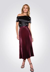 Top in satin nero scollato sulle spalle abbinato a una gonna midi plissettata bordeaux. Le calzature includono sandali neri con cinturini e dettagli in strass.