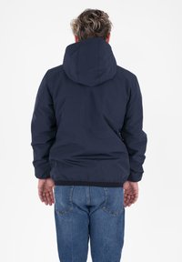 Persoon die met de rug naar de camera staat, gekleed in een marineblauwe hoodie en blauwe jeans, tegen een effen witte achtergrond.