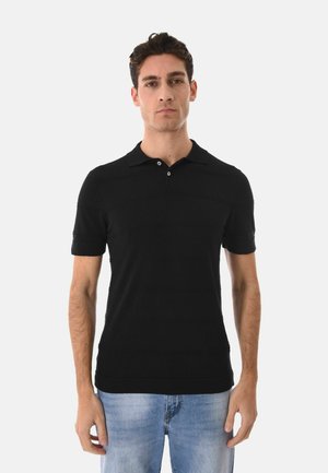 Uomo in piedi, rivolto in avanti, che indossa una polo nera a maniche corte e jeans azzurri chiari su sfondo bianco semplice.