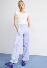 Ung person med kort blont hår står med händerna i fickorna på lila träningbyxor, iförd en figurnära vit topp och lila sportskor.