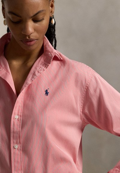 Γυναίκα που φοράει κόκκινο και λευκό ριγέ πουκάμισο με κουμπιά και ναυτικό σήμα κεντημένο, κοιτάζοντας προς τα κάτω σε απλό γκρι φόντο.