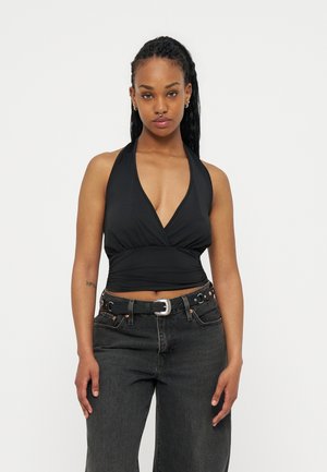 Γυναίκα με μακριά πλεξούδα, φορώντας μαύρο τοπ με τιράντες στον λαιμό και σκούρο τζιν ψηλόμεσο με μαύρη ζώνη, πάνω σε λευκό φόντο.