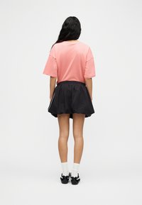Femme aux longs cheveux foncés vue de dos, portant un t-shirt corail oversize, une jupe plissée noire avec des bordures blanches, des chaussettes blanches et des baskets noires.
