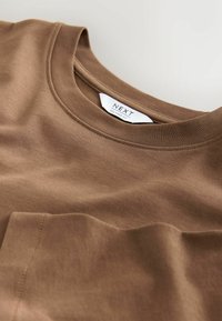 Tricou din bumbac maro, cu o croială relaxată, guler rotund și un buzunar pe piept. Textură fină cu detalii subtile de cusătură.