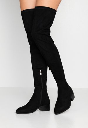 Bottes noires en daim au-dessus du genou avec talons blocs bas et fermetures éclair latérales, portées par une personne sur fond blanc.