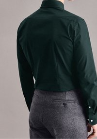 Osoba ubrana w ciemnozieloną koszulę z długim rękawem i szare spodnie wizytowe, pokazana z tyłu na neutralnym tle.