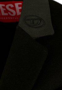 Cappotto di lana nero con colletto strutturato, logo ricamato sul colletto e etichetta rossa a contrasto all'interno. Tessuto morbido e strutturato.