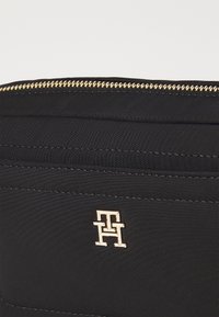 Wnętrze czarnej torby materiałowej z złotym zamkiem błyskawicznym i złotym emblematem logo o geometrycznym wzorze. Zawiera kieszenie na karty i teksturowane przeszycia.