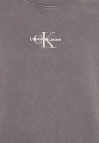 Felpa grigia con collo a girocostina. Presenta un logo bianco ricamato: "Calvin Klein Jeans" con "CK" in evidenza. Texture morbida.