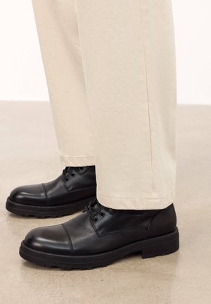 Cizme negre din piele până la gleznă, cu vârful rotund, dotate cu șireturi, o parte superioară texturată și o talpă groasă din cauciuc, purtate cu pantaloni bej, drepți.