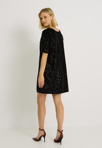 Robe noire en sequins avec une coupe ample et des manches courtes, dotée d'un col rond et d'une longueur au-dessus du genou. Portée avec des talons hauts marron.