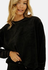 Jeune femme avec des cheveux blonds ondulés portant un haut noir à manches longues ample et un pantalon noir, souriant avec une main près de sa taille.