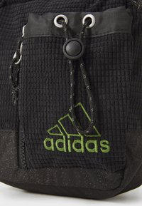 Mochila de tela texturizada negra con un prominente logo de Adidas bordado en verde, que cuenta con un cierre de cordón y detalles en metal plateado.
