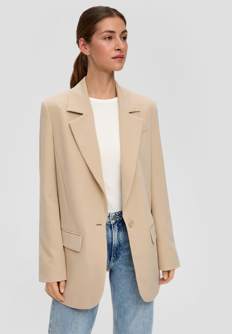 Beiger, taillierter Blazer mit Reverskragen, einzelner Knopfverschluss und zwei vorderen Taschen; kombiniert mit einem weißen Oberteil und blauen Jeans.