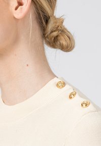 Profil d'une femme blonde avec les cheveux en chignon bas, portant un pull crème côtelé avec trois boutons décoratifs dorés sur l'épaule.