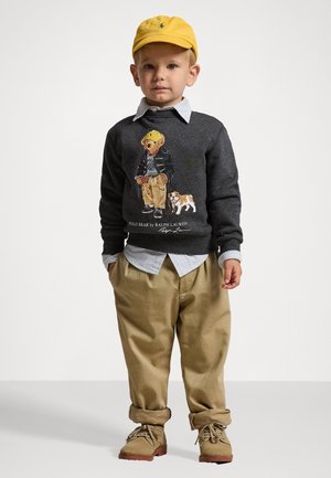 Suéter gris con gráfico de oso, superpuesto a una camisa de cuello azul claro. Pantalones caquis y zapatos beige, rematados con una gorra amarilla. Atuendo casual.