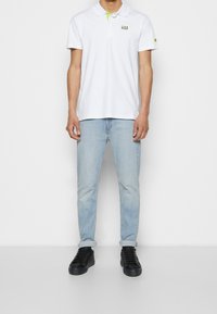 Biała koszulka polo z zielonym akcentem przy kołnierzyku, logo na piersi, zestawiona z jasnoniebieskimi dżinsami i czarnymi trampkami.