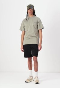 Camiseta de algodón verde oliva de gran tamaño, pantalones cortos negros, calcetas deportivas blancas, zapatillas beiges y una gorra de camuflaje con un detalle de logo.