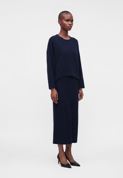 Femme debout portant un haut à manches longues bleu marine assorti et une jupe midi avec des talons pointus noirs sur un fond uni.