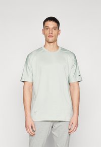 Camiseta de manga corta verde claro con cuello redondo, con mangas raglán y un pequeño logo en la manga izquierda. Textura de tela suave.