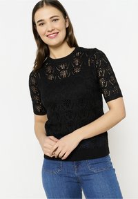 Juodas megztinis trumpomis rankovėmis, pasižymintis atvirais nėrinių raštais, grioveliu iškirptėje ir apačioje, derinamas su mėlynomis džinsomis. Audinys atrodo lengvas.