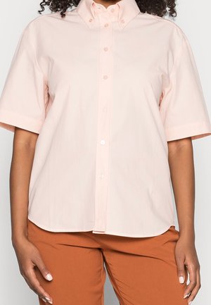 Person iført en lyserød kortærmet skjorte med knapper, kombineret med rustfarvede bukser, stående foran en ensfarvet baggrund.