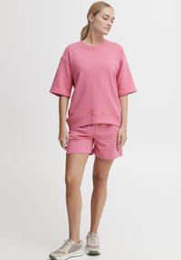 Rosa bomulls oversized sweatshirt med korte ermer, kombinert med matchende shorts. Skoene er lys grå med mørkegrønne detaljer og teksturerte såler.
