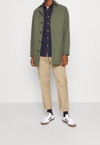 Manteau vert olive avec fermeture à boutons, chemise à carreaux navy en dessous, pantalon beige et baskets blanches avec rayures noires. Design simple.