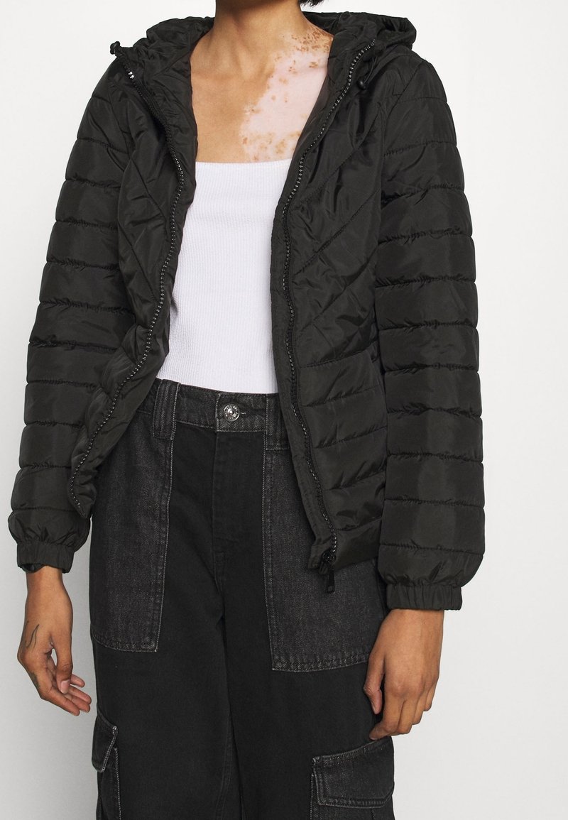 Personne portant une veste matelassée noire sur un débardeur blanc et un pantalon noir taille haute avec de grandes poches avant, debout les bras détendus.