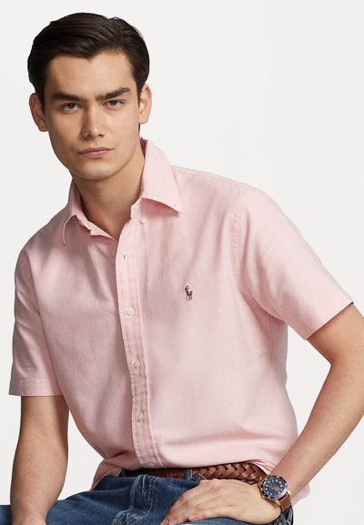 Kortermet rosa skjort med knapper og et lite brodert logo, kombinert med jeans, som viser en jevn stofftekstur og klassisk kragedesign.