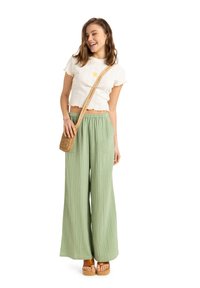 Weißes geripptes Croptop mit floraler Stickerei, kombiniert mit hellgrünen, weit geschnittenen Hosen. Das Model hält eine geflochtene Umhängetasche und trägt Plateausandalen.