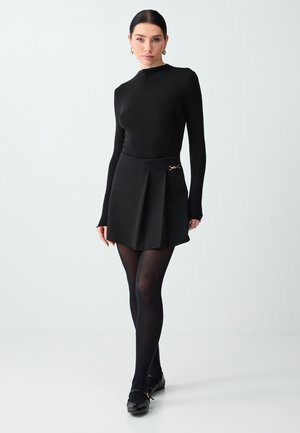 Femme aux cheveux foncés portant un haut noir à manches longues, une jupe plissée noire avec un détail en chaîne dorée, des collants noirs et des chaussures noires, sur un fond uni.