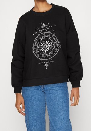 Persoon draagt een zwarte trui met een witte zon- en oogafbeelding en de tekst "Mijn kroon rechtzetten, mijn eigen toekomst maken," gecombineerd met een blauwe spijkerbroek.