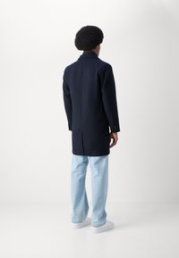 Abrigo navy con un corte ajustado, abertura sencilla en la parte posterior y puños abotonados, combinado con jeans azul claro y zapatillas blancas.