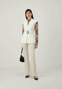 Nainen, joka on pukeutunut valkoiseen vyölliseen liiviin leopardikuosisen pitkähihaisen paidan päälle, beigeihin kuvioituihin leveälahkeisiin housuihin, mustiin kenkiin ja joka pitää käsissään pientä mustaa käsilaukkua.