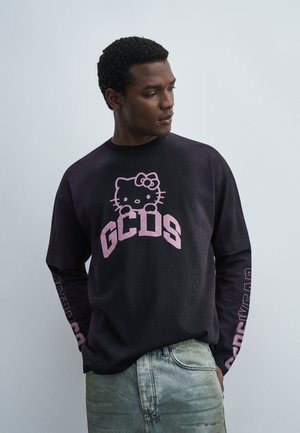 Hombre con camiseta negra de manga larga con Hello Kitty rosa y logo de GCDS, combinado con vaqueros azul claro, mirando hacia un lado sobre un fondo liso.