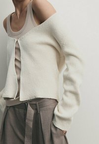 Cremefarbener, gestrickter Cardigan mit cropped Design, langen Ärmeln und Knopfverschluss. Getragen über einem beigen Tanktop und braunen Hosen.