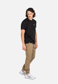 Mann mit lockigem Haar, der ein schwarzes T-Shirt, beige Jogginghosen und weiße Turnschuhe trägt, steht und schaut nach unten vor weißem Hintergrund.