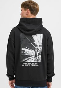 Zwarte hoodie met een grote grafische print van een zwart-wit stedelijk landschap op de achterkant. Zacht materiaal, ontspannen pasvorm, geribbelde manchetten.