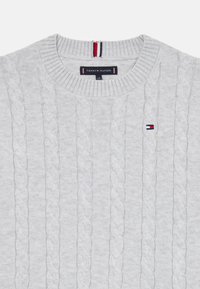Pull en maille torsadée gris clair avec un col rond côtelé et un petit patch logo avec des accents rouge, blanc et bleu. Motif texturé sur l'ensemble.
