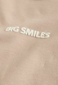 Tissu beige avec texte blanc en relief indiquant « BIG SMILES » incliné sur la surface.