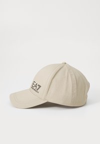 Căciulă de baseball din bumbac bej cu o margine curbă, având un logo negru pe partea din față și detalii de cusătură. Vedere laterală care arată forma rotunjită.