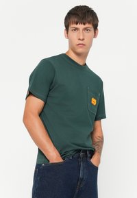 Camiseta de algodón verde con mangas cortas, cuello redondo, bolsillo frontal y detalle de etiqueta naranja. Presenta costuras visibles y un ajuste relajado.