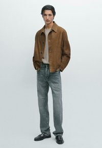 Brun mockajacka med krage, beige tröja under, ljusgrå raka jeans och svarta loafers. Enkel, minimalistisk design.