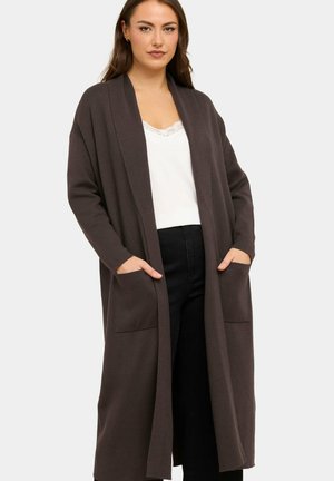Frau trägt einen langen dunkelbraunen offenen Cardigan mit Taschen, ein weißes Spitzen-Top und schwarze Hosen, steht vor einem schlichten Hintergrund.