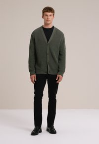 Zelen rebrast kardigan z V-izrezom in gumbi, nošen čez črno srajco, v kombinaciji s črnimi hlačami in čevlji, na nevtralnem ozadju.