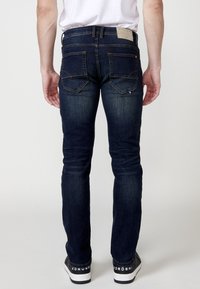 Jeans in denim blu scuro con design aderente, cinque tasche, cuciture a contrasto e etichetta sulla vita. Indossati con sneakers nere.