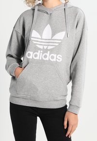 Šedá mikina s kapucí vyrobená z měkké tkaniny, s velkým bílým logem Adidas a třemi bílými pruhy. Obsahuje přední kapsu a kapuci se šňůrkami.