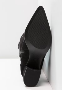 Musta mokkanahkasaappaat, joissa on terävä kärki ja teksturoitu kumipohja, aseteltuna valkoista seinää ja pintaa vasten.